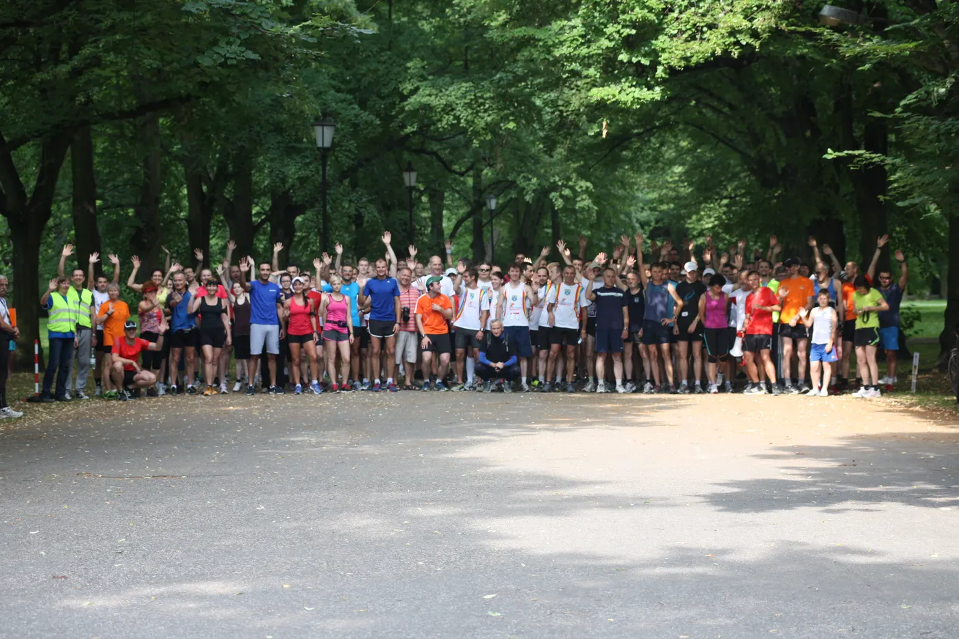 Kilkadziesiąt osób w strojach sportowych macha na starcie biegu parkrun w alejce parku w Łodzi